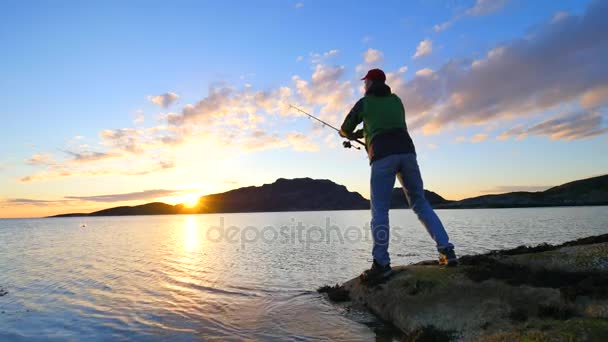 L'uomo attivo sta pescando in mare dalla costa rocciosa. Pescatore controllare spingendo esca sulla lenza, preparare canna e poi gettare esca in acqua pacifica. Silhouette da pescatore al tramonto — Video Stock