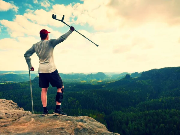Happy tourist with forearm crutch above head achieved mountain peak. Hiker with broken knee in immobilizer — Stock Photo, Image