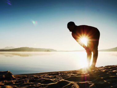Egzersiz ve göl kumsalda güneş doğarken germe etkin adam silüeti.