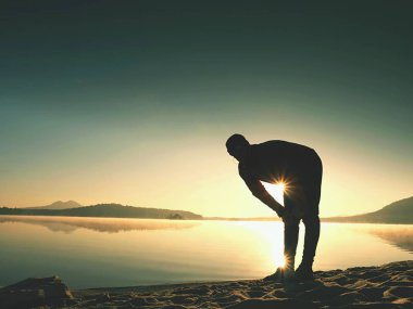 Egzersiz ve göl kumsalda güneş doğarken germe etkin adam silüeti.