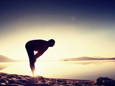 Egzersiz ve göl kumsalda güneş doğarken germe etkin adam silüeti.