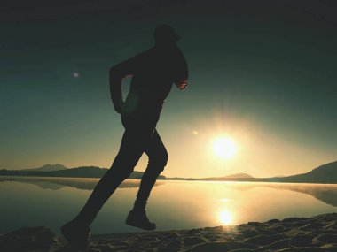 Egzersiz ve göl kumsalda güneş doğarken germe etkin adam silüeti.