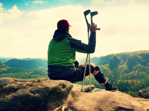 Уставший турист сидит с поврежденным коленом в иммобилайзере или камне, держит медицинский столб. Открытый лесной ландшафт — стоковое фото