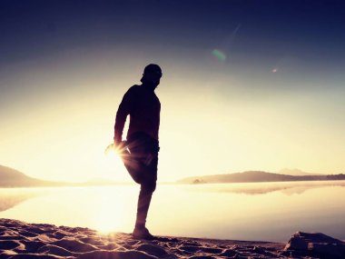 Egzersiz ve göl kumsalda güneş doğarken germe etkin adam silüeti.
