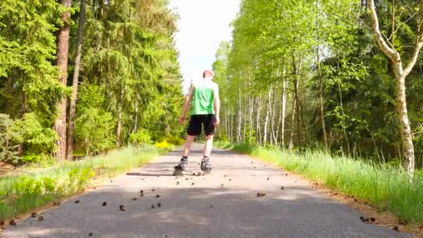 Vista trasera al patinador en línea en singlet verde. Patinaje en línea al aire libre sobre asfalto liso en el bosque. Hombre de piel clara patear en conos de pino en el camino, moviéndose con el centro de gravedad . — Vídeo de stock