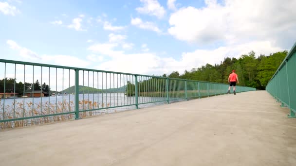 Вид сзади на фигуриста в красной футболке и черных брюках на мосту. Открытый каток на гладкой бетонной площадке на озерном мосту. Светлый человек в сапогах на колесах — стоковое видео