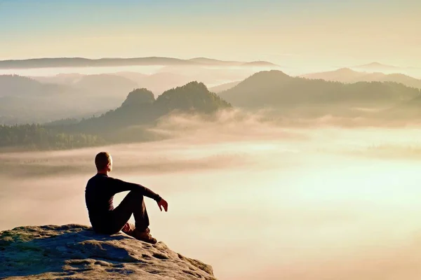 Hiker in black on the rocky peak. Wonderful daybreak in mountains, heavy orange mist in valley. Man sit on the rock. — Stock Photo, Image
