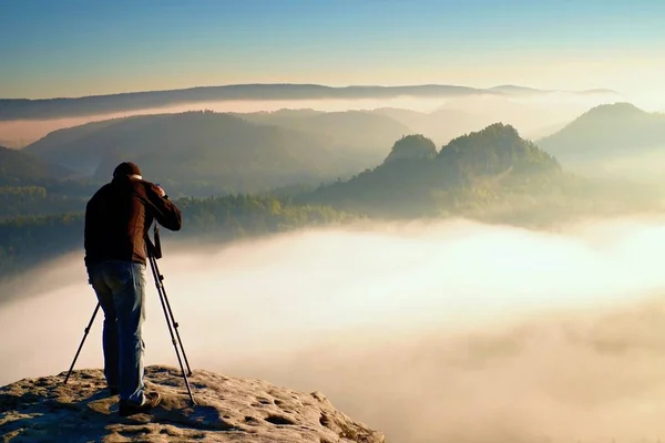 Profi an der Klippe. Naturfotograf fotografiert mit Spiegelkamera auf Felsen. verträumte nebelige Landschaft, darunter orange-rosa Nebel. — Stockfoto