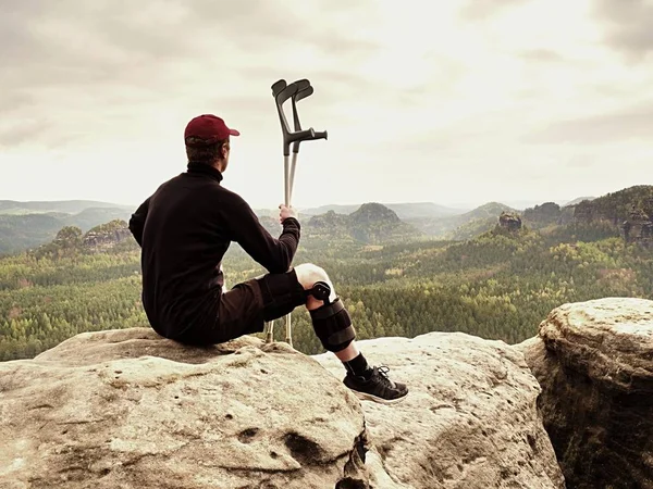 Tired hurt tourist with medicine crutches. Man with  broken leg in knee brace features resting on  exposed rocky summit