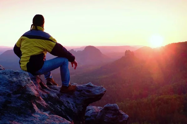 Vue arrière du randonneur masculin en veste jaune noire assis sur une falaise rocheuse tout en profitant du lever du jour au-dessus de la vallée — Photo