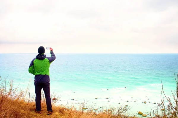 Man in green waterproof jacket takes photo of coastliny by smart phone. Cold windy day at sea.