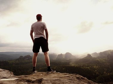 Man in t-shirt and pants. Tourist on mountain peak looking into landscape. clipart