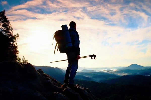 Tourist guide with pole in hand. Hiker with sporty backpack stand on rocky view point above misty valley. Sunny spring daybreak — Stock Photo, Image