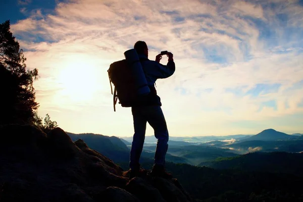 Turista com mochila pesada tira fotos com telefone inteligente. Homem no pico rochoso. Vale nebuloso sonhador abaixo — Fotografia de Stock