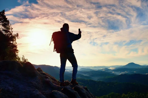 Турист с тяжелым рюкзаком фотографируется со смартфоном. Человек на скалистой вершине. Мечтательная туманная долина внизу — стоковое фото