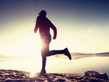 Egzersiz ve göl kumsalda güneş doğarken germe etkin adam silüeti.