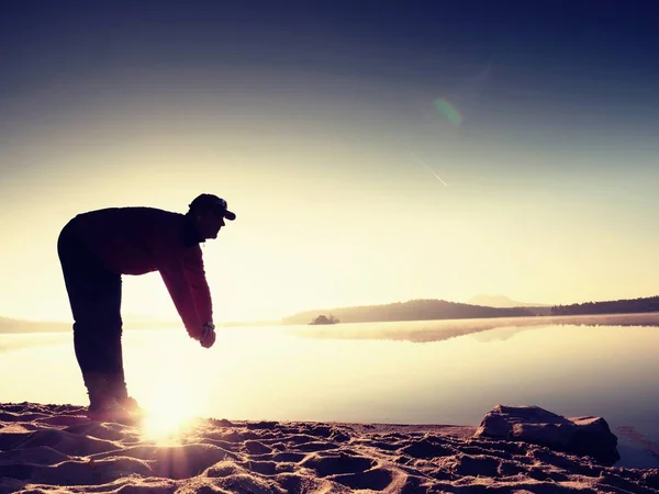 Egzersiz ve göl kumsalda güneş doğarken germe etkin adam silüeti. — Stok fotoğraf