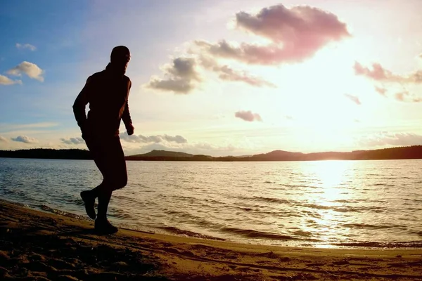 Uomo alto con occhiali da sole e cappello scuro è in esecuzione sulla spiaggia al tramonto autunnale — Foto Stock