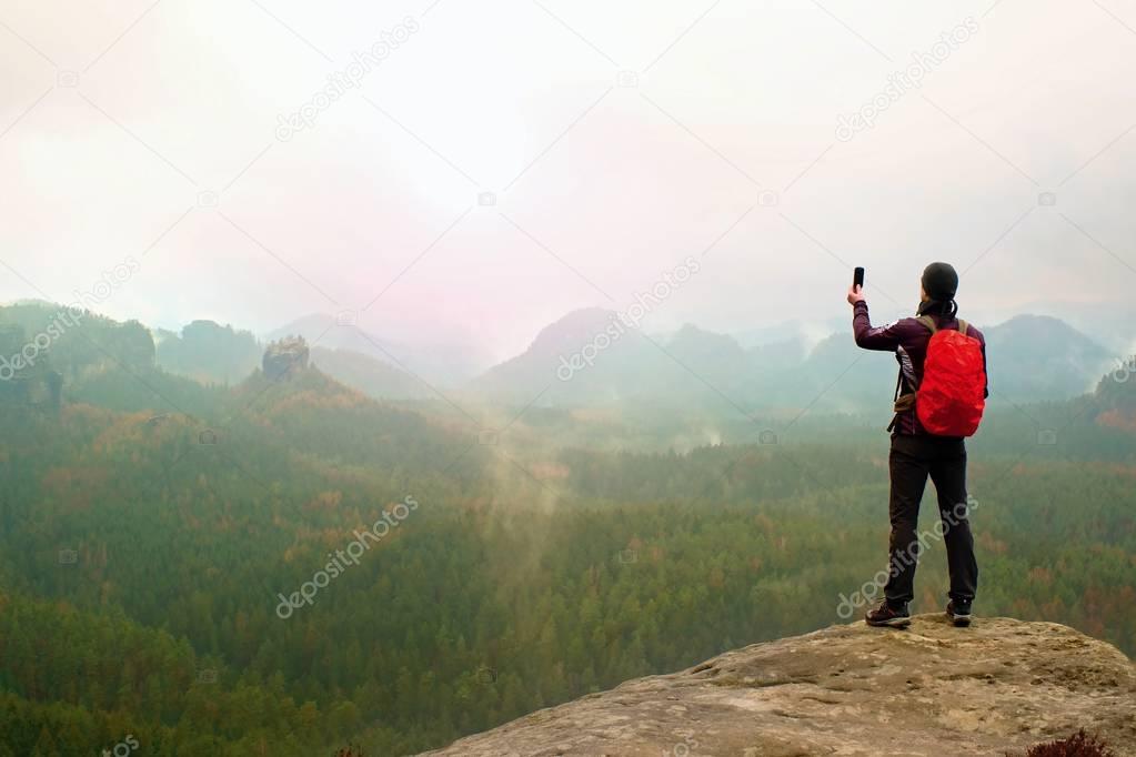 Tourist with backpack takes photos with smart phone of rainy vally. Dreamy foggy valley below