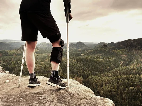 의학 목 발과 상처 관광 피곤. 무릎 중괄호 기능 노출된 바위 정상에 휴식에 부러진된 다리를 가진 남자 — 스톡 사진
