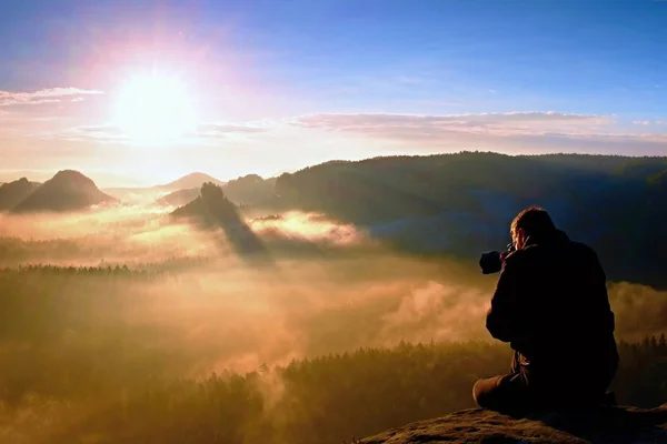 Turista feliz con cámara en las manos sentarse en el pico de roca arenisca y ver en la niebla de colores y niebla en el valle de la mañana . —  Fotos de Stock