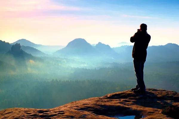 Professionelle Fotograf macht Fotos mit großer Kamera auf Fels. Gefährliche Lage am Ende der Klippe. verträumte Nebellandschaft, heiße Sonne darüber — Stockfoto