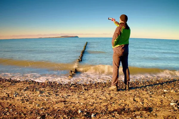 Turista mantenga el teléfono celular, tomar fotos de paisajes marinos de otoño en el rompeolas. Efecto vívido y vignetting . — Foto de Stock