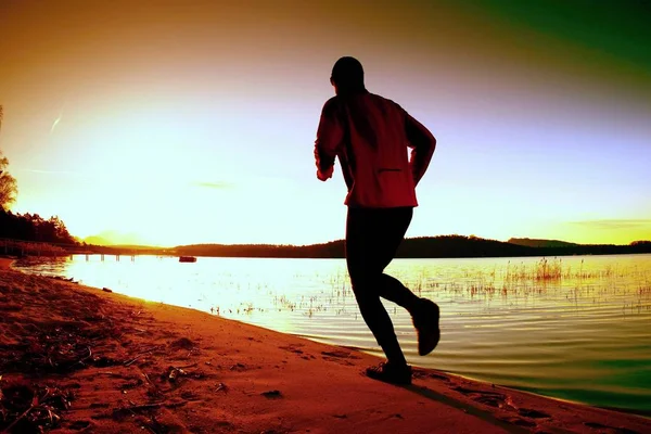 Hombre alto con cazador de viento rosa y gorra oscura correr en la playa al atardecer — Foto de Stock