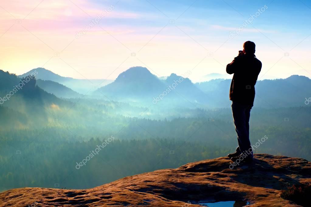 Professional photographer takes photos with big camera on rock. Dangerous possition at end of cliff.  Dreamy  foggy landscape, hot Sun above