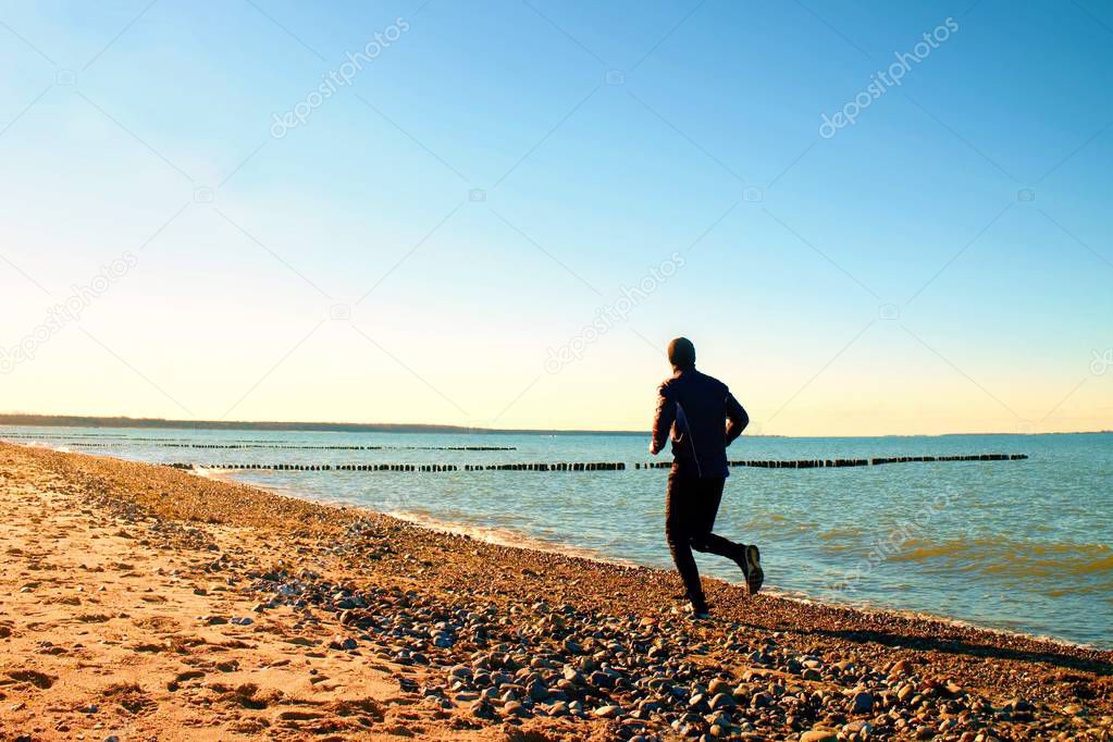 Tall man in dark sportswer running and exercising on stony beach at breakwater.