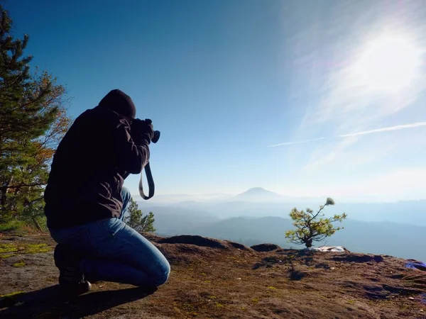 Fotógrafo para baixo de joelhos tira fotos com câmera de espelho no pico da rocha — Fotografia de Stock