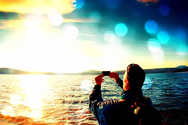 Grano de película. Cabello alto jengibre hombre turista tomando fotos o vídeo con teléfono móvil. Otoño día soleado en la costa del mar — Foto de Stock