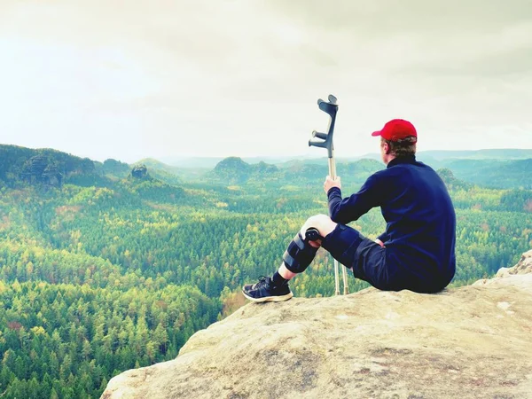 Tıp koltuk değneği ile yorgun yaralı turist. Kırık bacak diz küme ayracı özellikleri üzerinde dinlenme adamla kayalık Zirvesi maruz