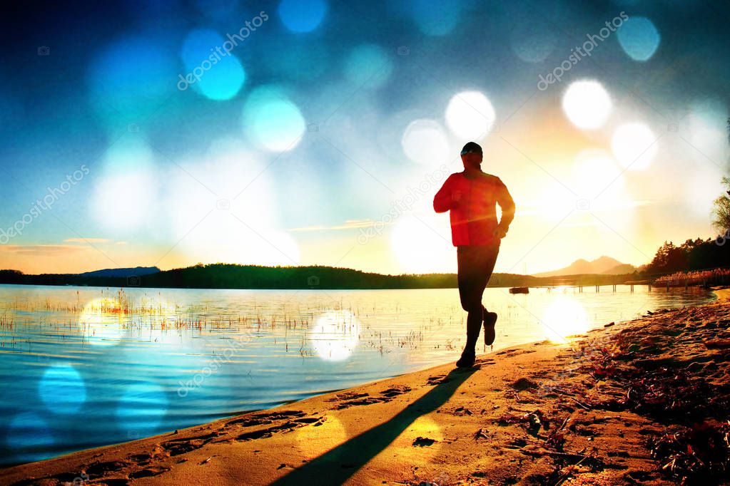 Film grain. Silhouette of sport active adult man running and exercising on the beach. Calm water, island and sunset sky background.