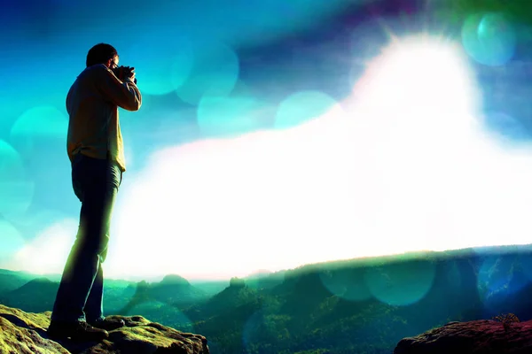 Film grain. Professional photographer takes photos with big camera on peak of rock. Dreamy misty landscape, hot Sun above — Stock Photo, Image