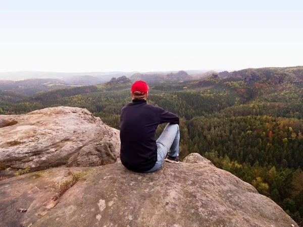 Caminante en Cliff. Turista en gorra roja, sudadera negra y jeans se sientan en un acantilado de montaña sobre el valle — Foto de Stock