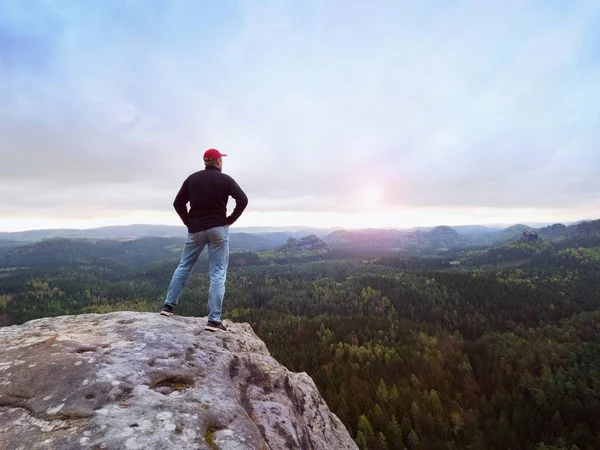 Hombre en jeans, sudadera y gorra roja en un acantilado rocoso agrietado. Día melancólico en las montañas de arenisca, valle del bosque profundo —  Fotos de Stock