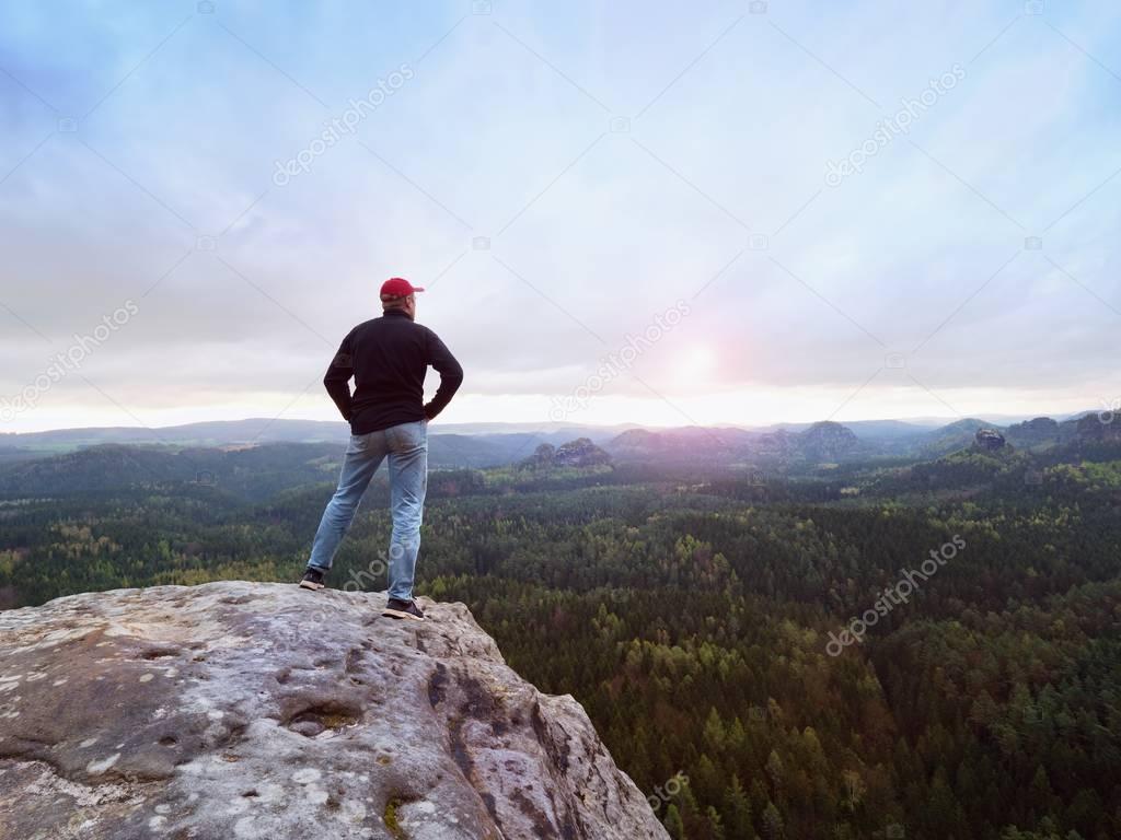Man in jeans, sweatshirt and red cap on cracked rocky cliff.  Melancholy day in sandstone mountains, deep forest valley
