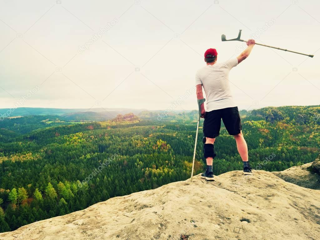 Happy man hiker holding medicine crutch above head, injured knee fixed in knee brace feature