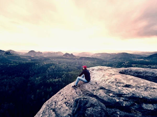 Caminante en Cliff. Turista en gorra roja, sudadera negra y jeans se sientan en un acantilado de montaña sobre el valle — Foto de Stock