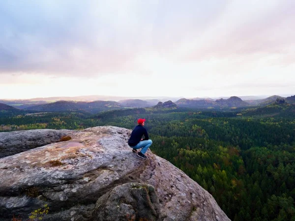 Wanderer auf Klippe. Tourist in roter Mütze, schwarzem Sweatshirt und Jeans sitzt auf scharfer Bergklippe über dem Tal — Stockfoto