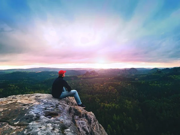 Wanderer auf Klippe. Tourist in roter Mütze, schwarzem Sweatshirt und Jeans sitzt auf scharfer Bergklippe über dem Tal — Stockfoto