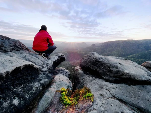 Volwassen toerist in zwarte broek, rode jas en donkere dop zitten op de rand van de klif en kijken naar de vallei — Stockfoto