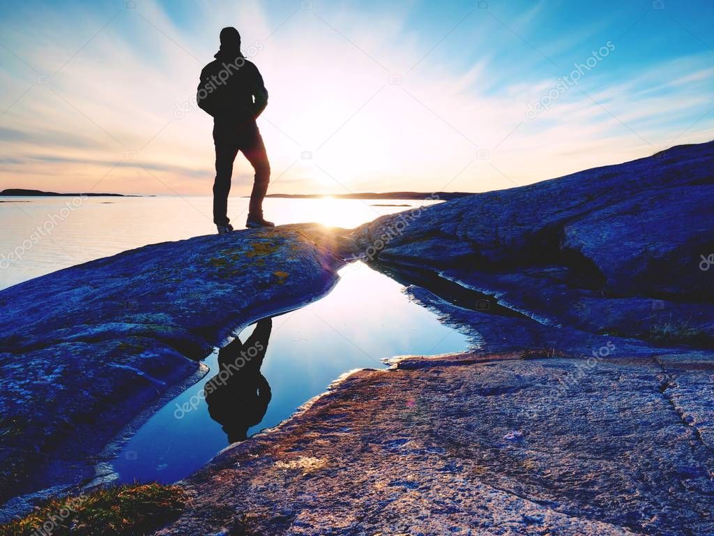 Hiker in dark sportswear with poles and sporty backpack. Coastline trail on rocky shore. Alone tourist enjoy