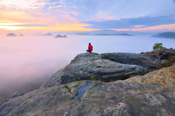 Hiker in squatting position on peak of rock and watching into colorful mist and fog in morning valley. — Stock Photo, Image