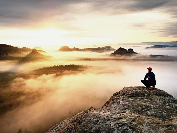 Erwachsene Touristen in schwarzer Hose, Jacke und dunkler Mütze sitzen am Rand der Klippe und blicken auf das neblige Hügelland. — Stockfoto