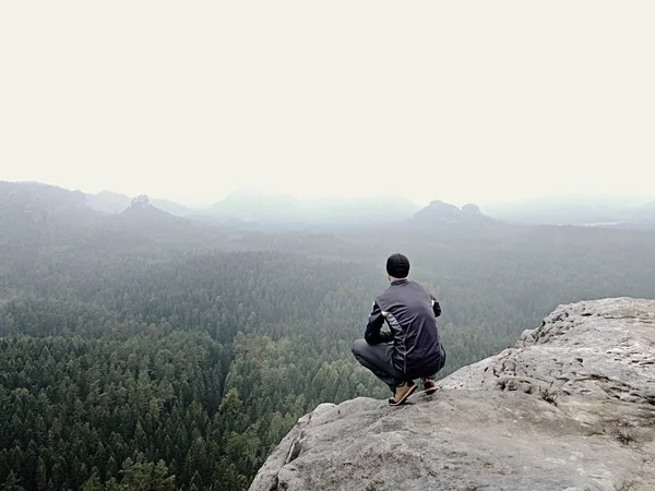 Vista trasera de excursionista solo en traje oscuro al aire libre sentarse en el borde. Pico rocoso agudo sobre el valle de las montañas —  Fotos de Stock