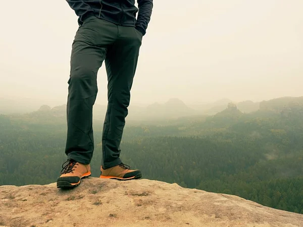 Pernas masculinas em calças de caminhada escuras e sapatos de trekking de couro no pico da rocha acima do vale nebuloso. Esboço da colina — Fotografia de Stock