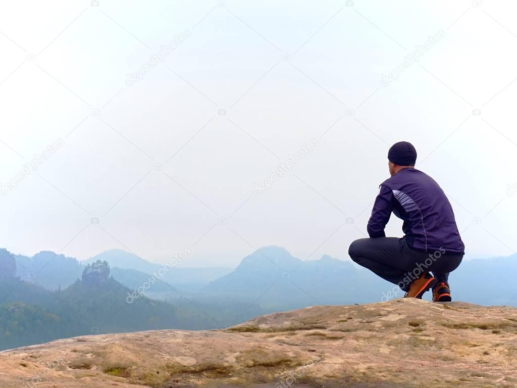 Adult tourist in black trousers, jacket and dark cap sit on cliff's edge and looking into mist