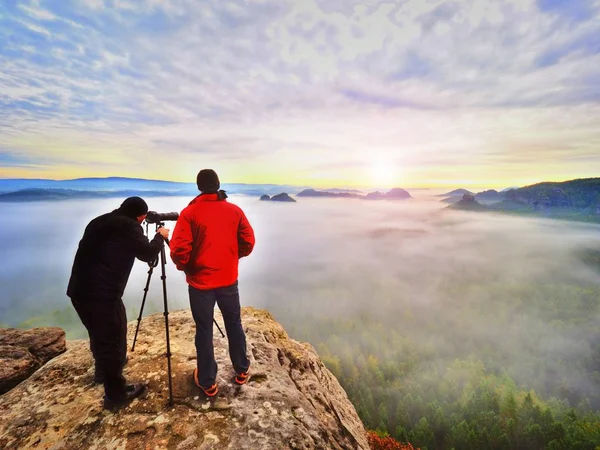 Fotograaf met oog op de zoeker van de camera op statief verblijf op klif en neemt foto's, praten vrienden. — Stockfoto